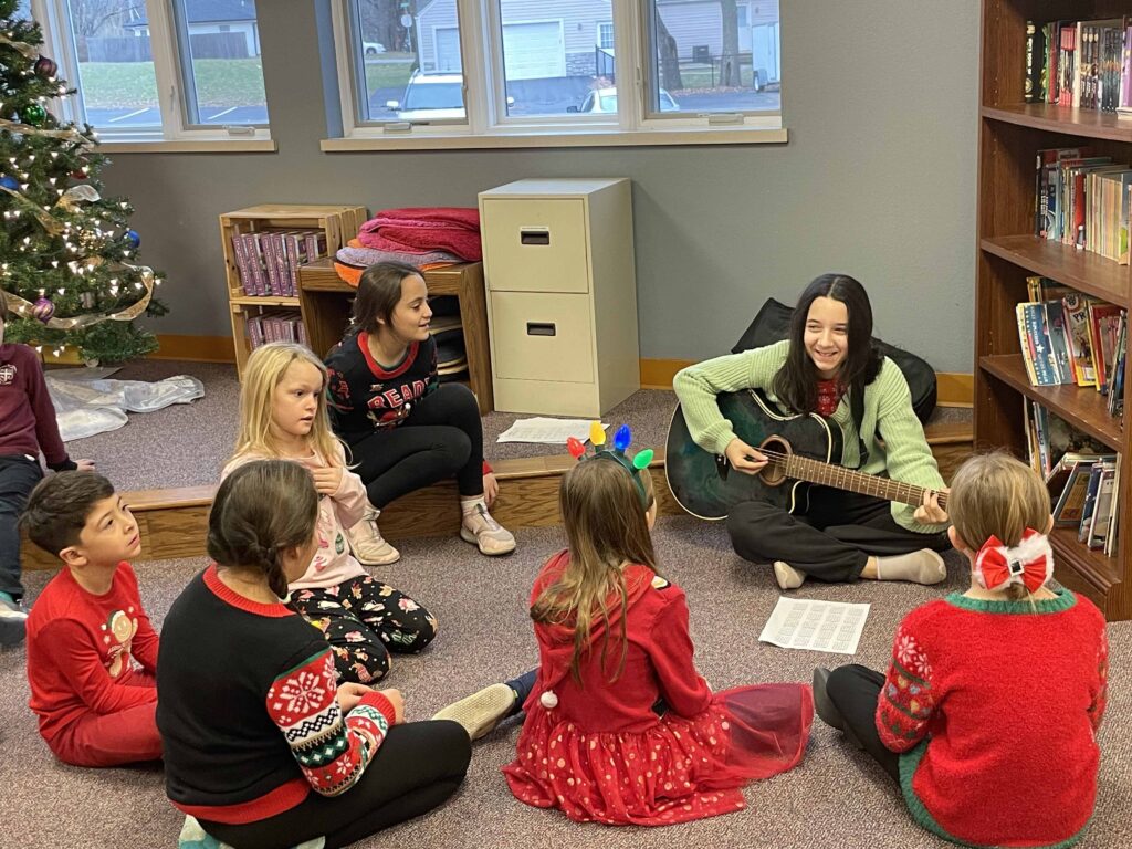 Young students sit in a circle at Christmas time while an elder student plays the guitar for them