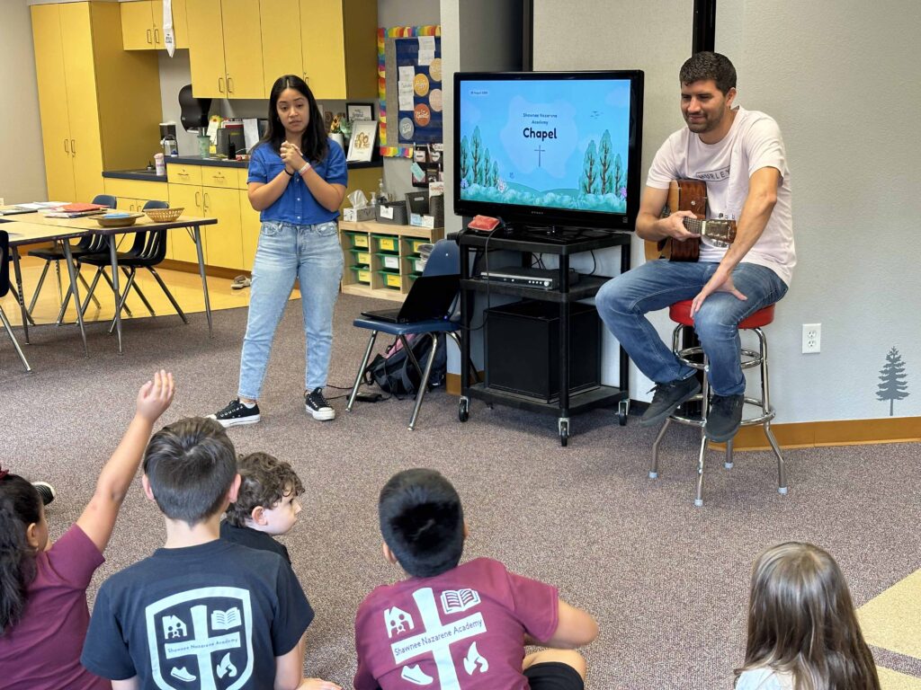 Young students listen to a chapel presentation with their Teacher who plays them an acoustic guitar